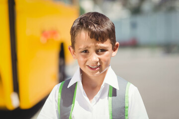 Asian child standing and waiting by yellow school bus on sunny pickup area with grey-green backpack