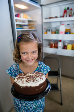 Naklejki Smiling girl in blue dress holding white-frosted chocolate cake near open refrigerator in kitchen
