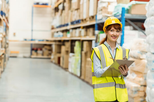 Woman in 30s wearing hard hat and vest checking clipboard pen near warehouse shelves, copy space