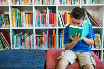 School-age boy sitting on blue and red bench reading green hardcover book in library, copy space