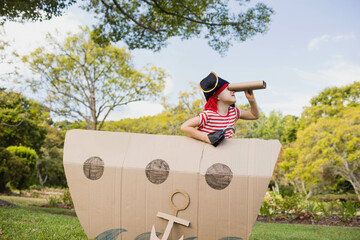 Child pirate peering through paper-tube telescope behind large cardboard ship cutout in sunny park