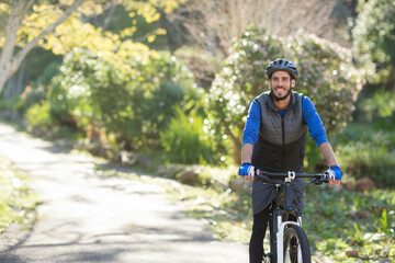 Naklejka premium Male cyclist riding mountain bike along sunlit paved park trail with helmet and gloves, copy space