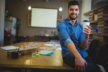 Man sitting on desk edge in office sipping coffee with blank projector screen behind, copy space