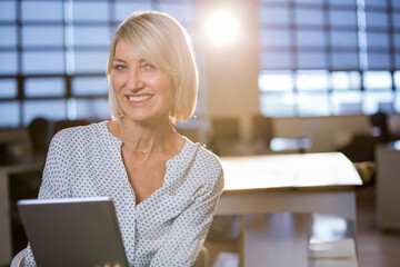 Businesswoman smiling while holding tablet at office desk in patterned blouse with conference table