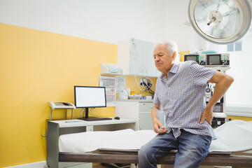 Senior man sitting on exam table in medical room with computer desk and monitors, copy space