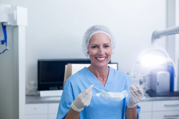 Smiling nurse wearing hairnet scrubs and latex gloves holding mask in clinic near monitor lamp