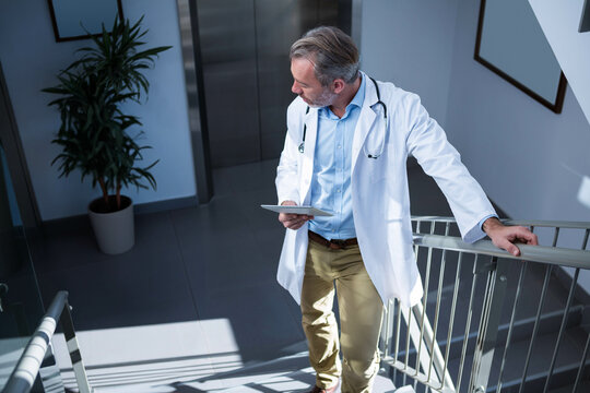 Doctor wearing lab coat and stethoscope, holding tablet leaning on railing near elevator and plant