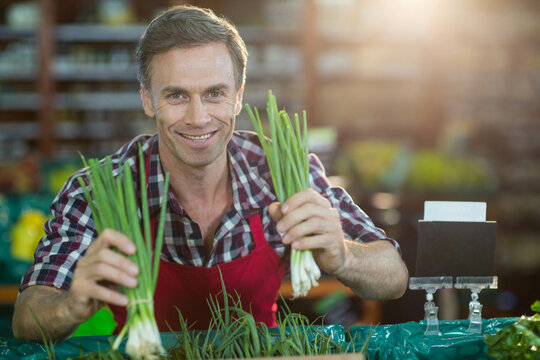 Male vendor holding up green onion bunches at grocery store produce table wearing red apron - Powered by Adobe