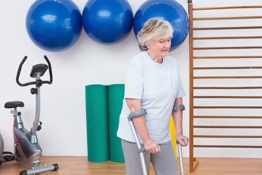 Senior White woman using forearm crutches balancing in rehab gym with balls and bike, copy space
