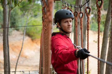 Man in mid thirties wearing helmet and harness, gripping cable and balancing on forest rope course