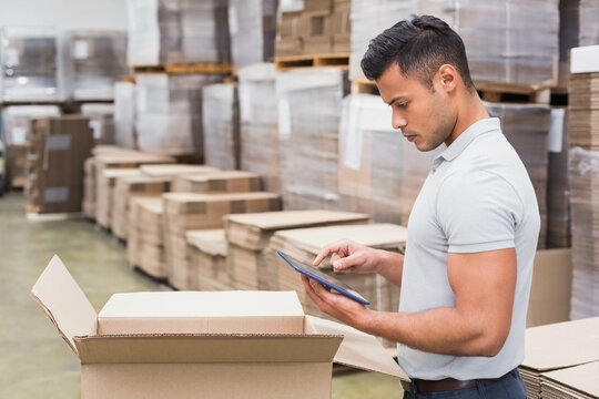 Male worker verifying inventory with tablet in warehouse next to open box, pallet stacks copy space