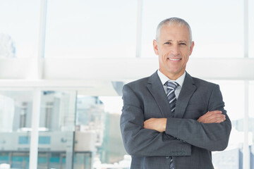 Senior man standing with arms crossed in corporate office by floor-to-ceiling windows, copy space