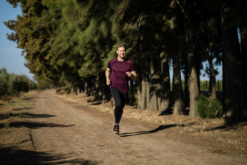 Male jogger running along dirt trail through pine forest wearing maroon athletic shirt