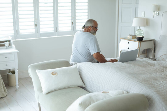 Senior man is working on laptop on bed in minimalist bedroom with white shutters and nightstands