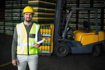 Male warehouse worker wearing helmet, vest holding clipboard near pallets and forklift, copy space