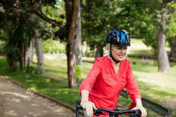 Obraz premium Woman riding bicycle along park dirt path wearing red shirt, helmet beside pond railing, copy space