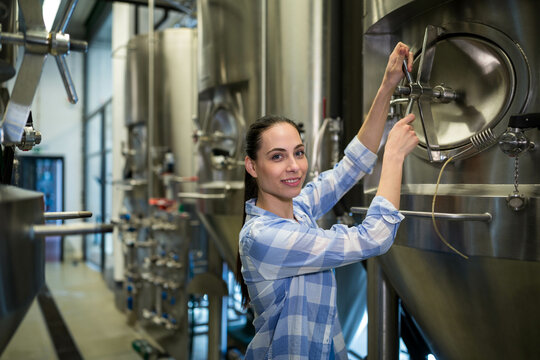 Female brewer adjusting latch on stainless steel fermentation tank amid valves in brewery facility