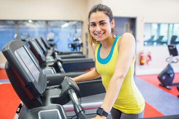 Fototapeta premium Hispanic woman in gym using treadmill, smiling, holding console, mirrored wall showing cardio bikes