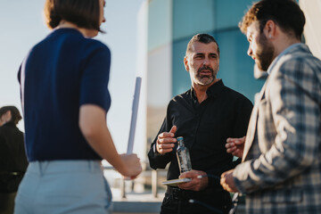 A group of diverse business people engage in conversation on a rooftop at sunset. They discuss...