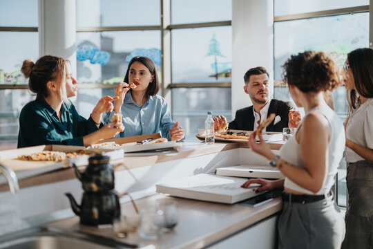 A group of colleagues take a break in the office kitchen, enjoying pizza and conversation. The atmosphere is relaxed and friendly, highlighting teamwork and collaboration in a modern work environment.