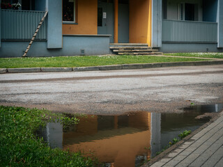 The entrance of a residential building with steps and balconies is reflected in a puddle on the...