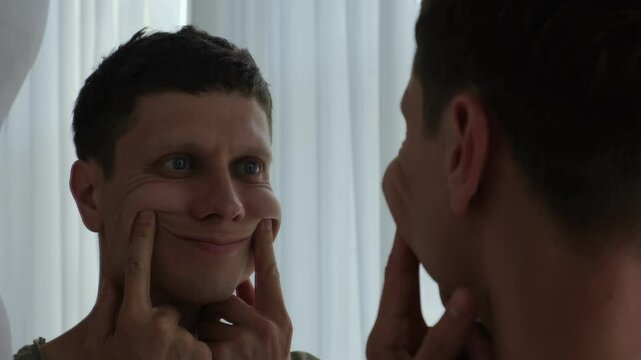 Depressed young man fakes a smile in the mirror. Psychological struggle, mental health concept.