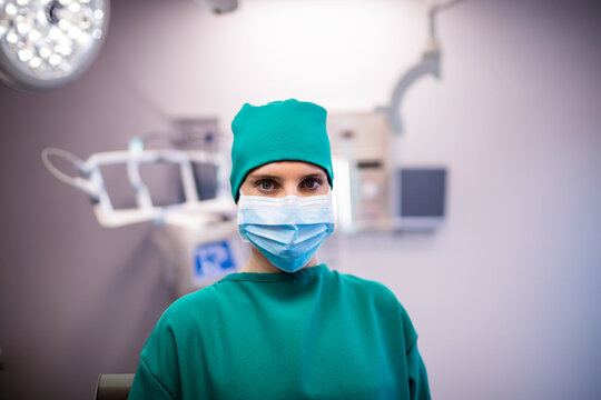 Female surgeon standing in green scrubs and mask amid surgical lights, monitor arms, medical panels