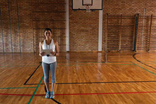 Fototapeta Instructor standing on polished court holding tablet and stopwatch under gym basketball hoop