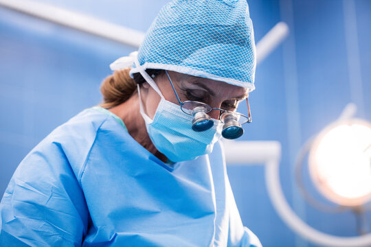 Senior surgeon leaning under operating room lights, wearing blue scrubs, cap, mask and loupes