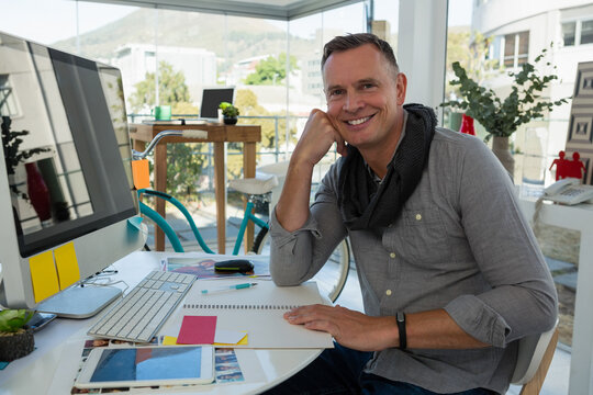 Mature man smiling while reviewing sketchbook near computer and sticky notes at office, copy space