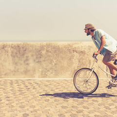 Man is cycling on white bicycle wearing straw hat along hexagonal stone promenade beside sea