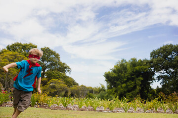 Boy wearing blue T-shirt showing red lightning bolt, red cape, eye mask running across grassy lawn