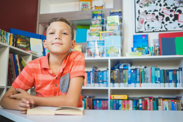 Boy reading open book at white table in school library reading corner with colorful books behind