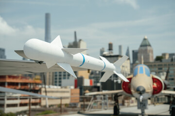 A white air-to-air missile is mounted under the wing of a military jet, photographed with an urban...