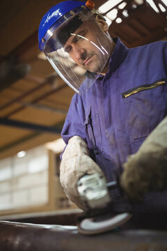 Male metalworker wearing helmet with face shield operating angle grinder on pipe at workbench