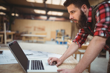 Craftsman leaning over table using laptop and reviewing blueprints under skylights in woodshop