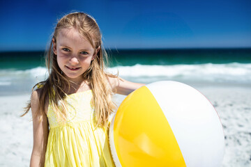Naklejka premium Child standing on white sand beach holding large yellow white inflatable beach ball, waves rolling