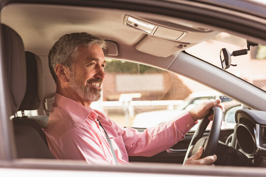 Man wearing pink shirt and seatbelt is smiling, gripping wheel in sunlit car near brick buildings