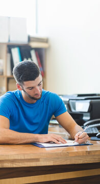 Clipboard holding papers while pen resting on wooden desk near bookshelf and printer by window