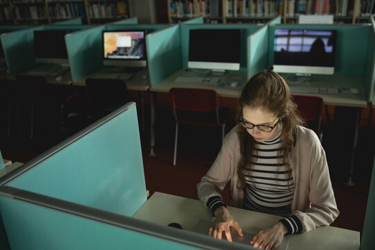 Teenage female student typing on laptop at library computer lab with teal desk dividers, copy space