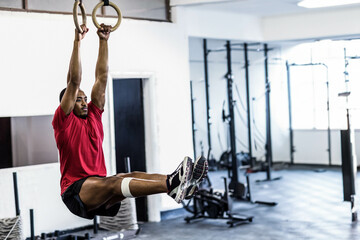 Man is performing hanging leg raise on wooden rings in gym, with knee tape and rigs