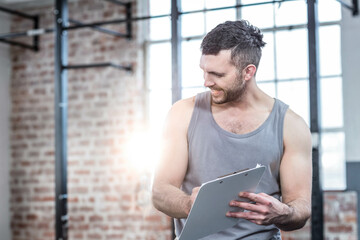 Man in gear holding clipboard reviewing inside gym with windows, pull-up bars and bench, copy space