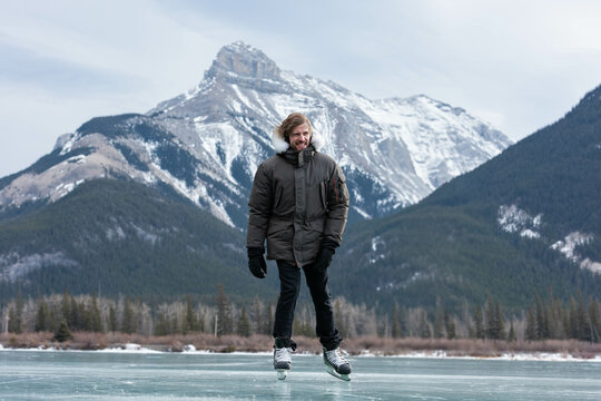 Lone man wearing winter jacket and gloves using ice skates on frozen lake near snowy peaks