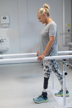 Senior woman practicing walking between parallel bars in rehabilitation clinic, with prosthetic leg