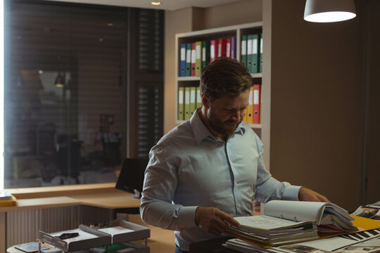 Middle aged man reviewing documents at office workspace with large binder and computer monitor