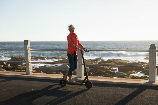 Woman standing on electric scooter at seafront promenade gripping handlebars near coastal guardrail