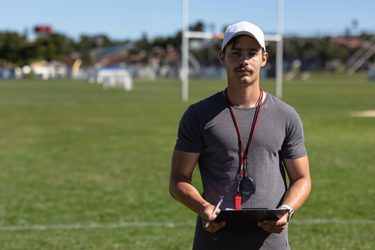 Male coach holding clipboard and pen while wearing baseball cap and whistle near rugby goal posts
