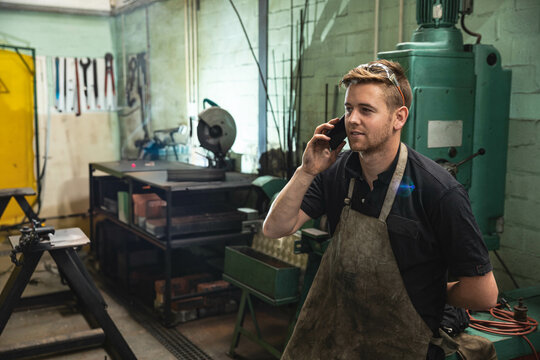 Male metalworker wearing apron examining smartphone and operating metal saw in workshop, copy space
