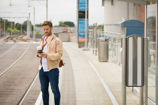 Thirty something man waiting on rail platform with satchel holding coffee cup and using smartphone