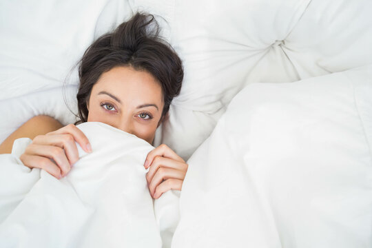 Woman gripping duvet and peeking over bed in bedroom with tufted headboard and white pillow